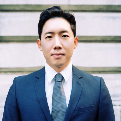 Jaden Hong in a navy suit, standing in front of a light stone wall.